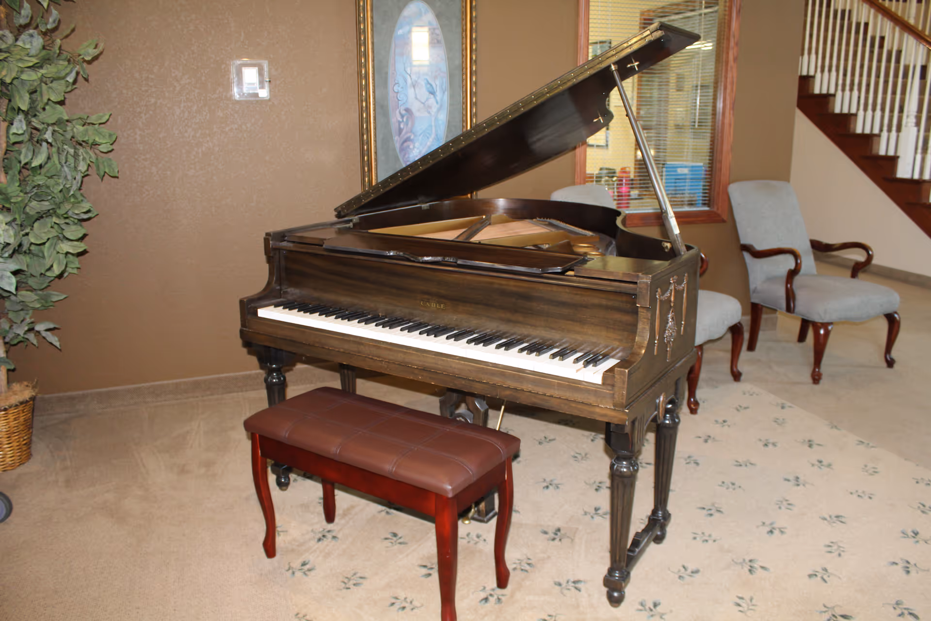 Interior room with a grand piano and a padded piano bench on a carpeted floor. Two upholstered chairs with wooden arms are positioned near a window with blinds. A potted plant is visible on the left side, and a staircase with white railings is in the background.