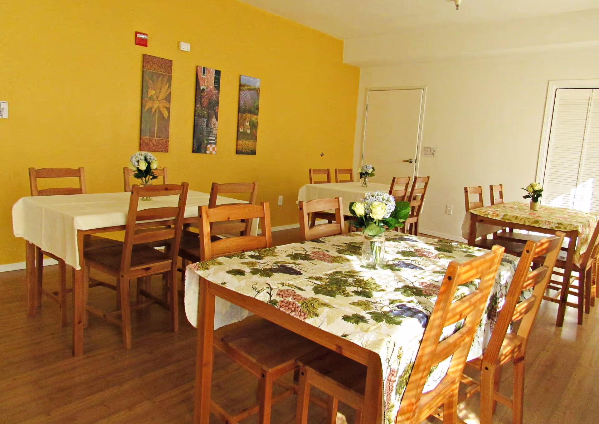 A dining room with several wooden tables and chairs. The tables are covered with tablecloths, some with floral patterns and others plain. Each table has a small vase with flowers. The room has a yellow accent wall with three vertical paintings and a wooden floor.