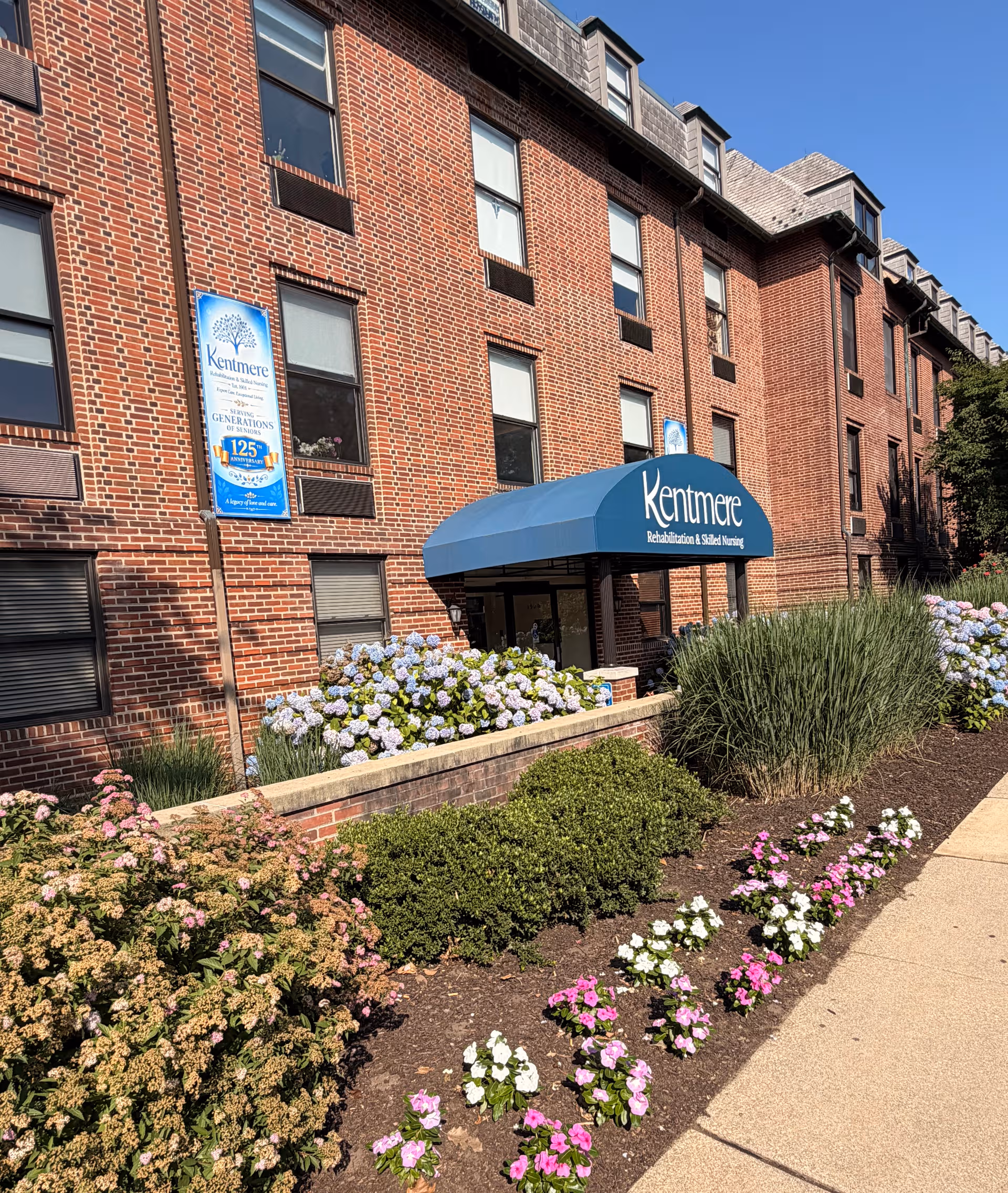 Brick senior living building with blue awning entrance and manicured flowerbeds along walkway.