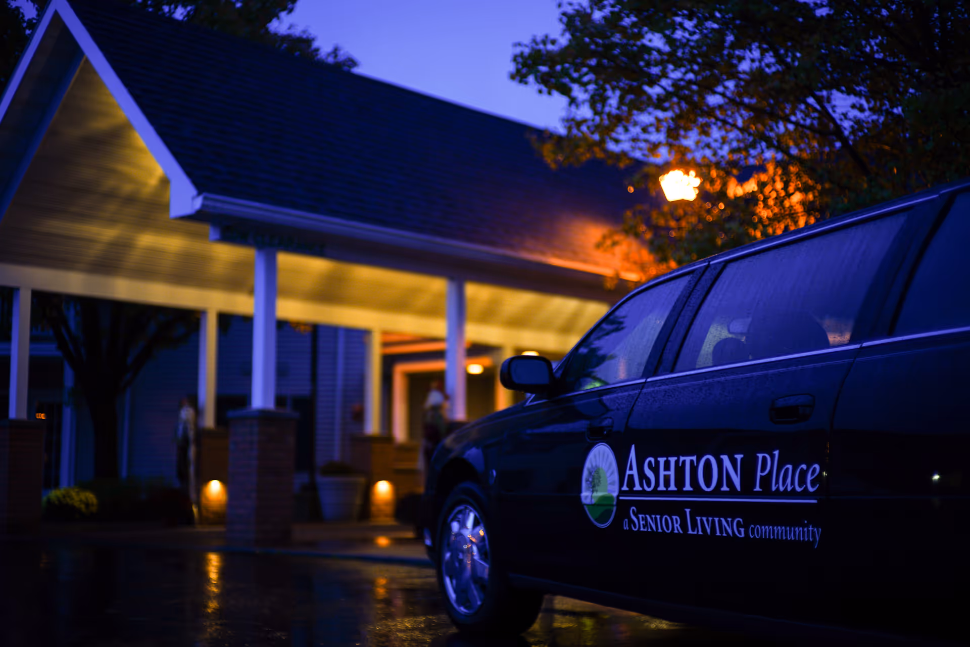 A dark-colored car with the logo and text 'Ashton Place a Senior Living community' parked outside a building with a covered entrance illuminated by warm lights during dusk or early evening.