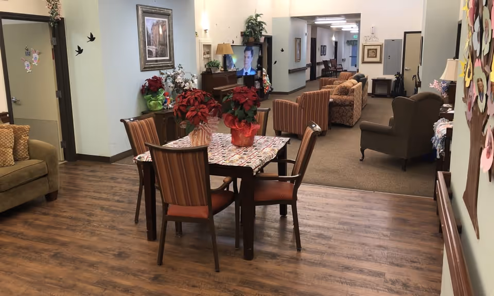 Interior view of a senior living facility common area with a wooden table and four striped chairs in the foreground. The table is decorated with a floral tablecloth and red poinsettia plants. To the left is a beige couch with patterned cushions. In the background, there is a hallway leading to more seating areas with armchairs and sofas. The walls are decorated with framed pictures and plants, and the floor transitions from wood to carpet.