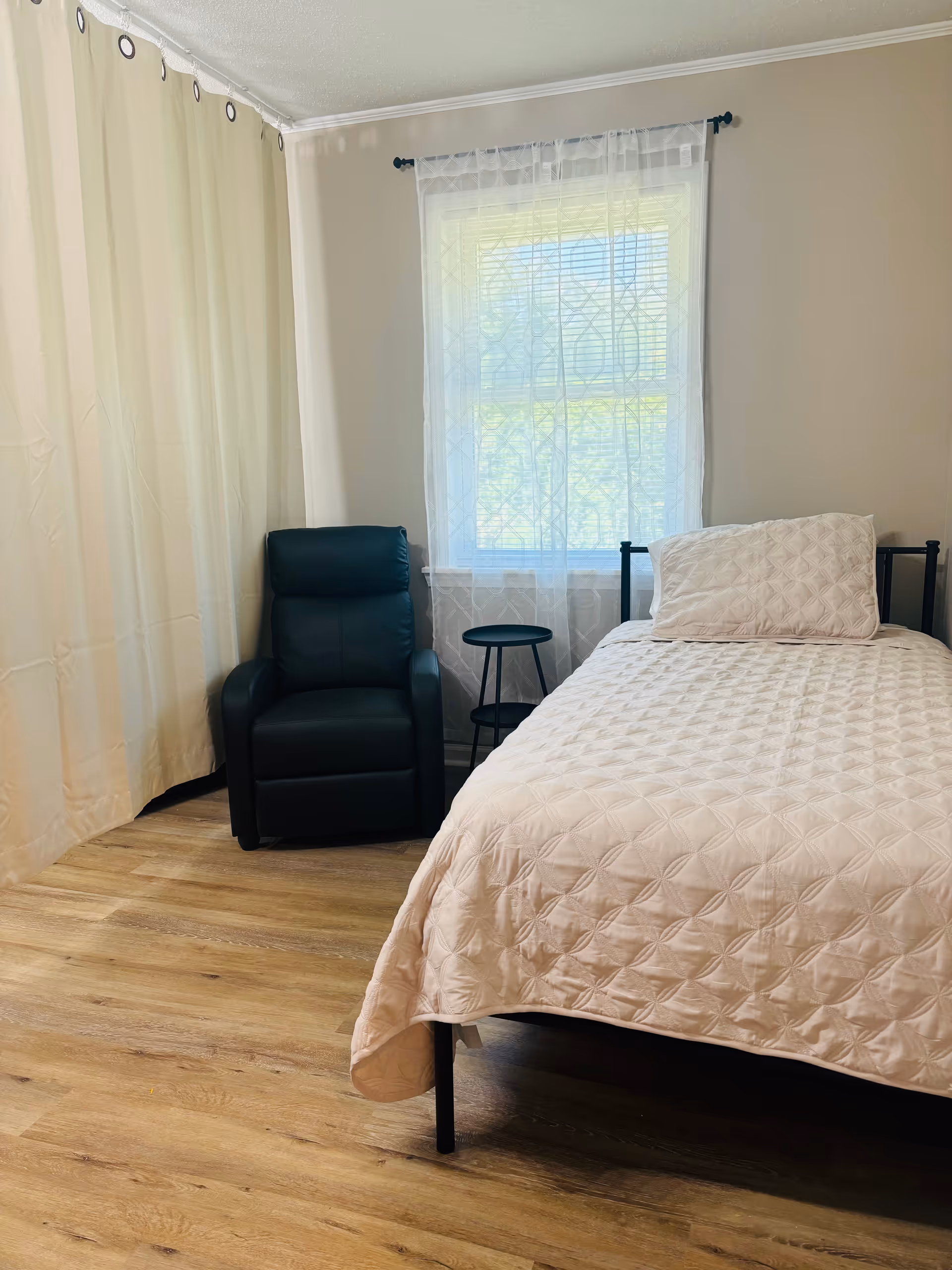 A small bedroom with a single bed covered in a light beige quilt and matching pillow. Next to the bed is a small black round side table and a black recliner chair. A window with sheer white curtains lets in natural light, and beige curtains cover the wall on the left side. The floor is wood with a light finish.