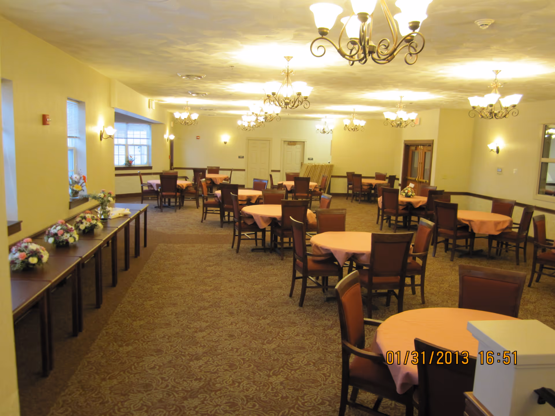 A spacious dining room with multiple round tables covered with pink tablecloths, each surrounded by several chairs. The room is well-lit with multiple chandeliers and wall sconces. There are flower arrangements on a long table along the left wall and on some dining tables. The carpet has a patterned design, and the walls are painted a light yellow color.