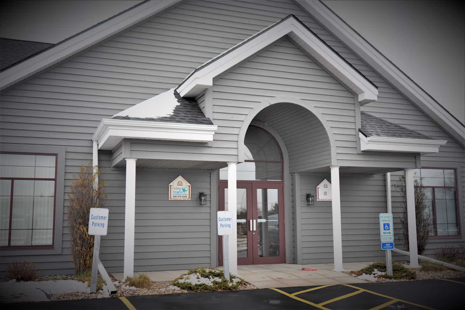 Gray senior living facility entrance with arched doorway and parking signs.