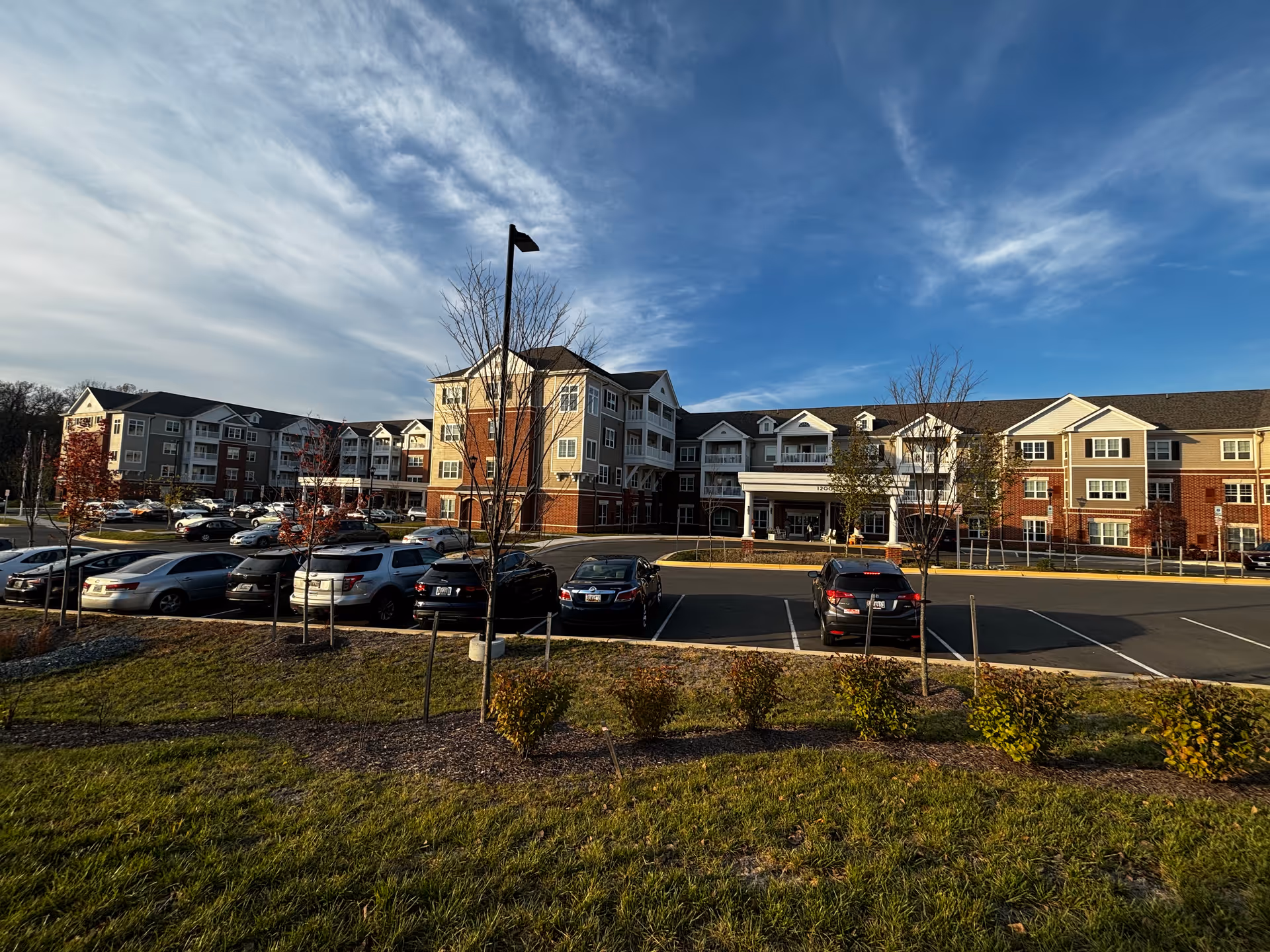 Front view of a multi-story senior living building with cars parked in a lot and a grassy foreground under a blue sky.