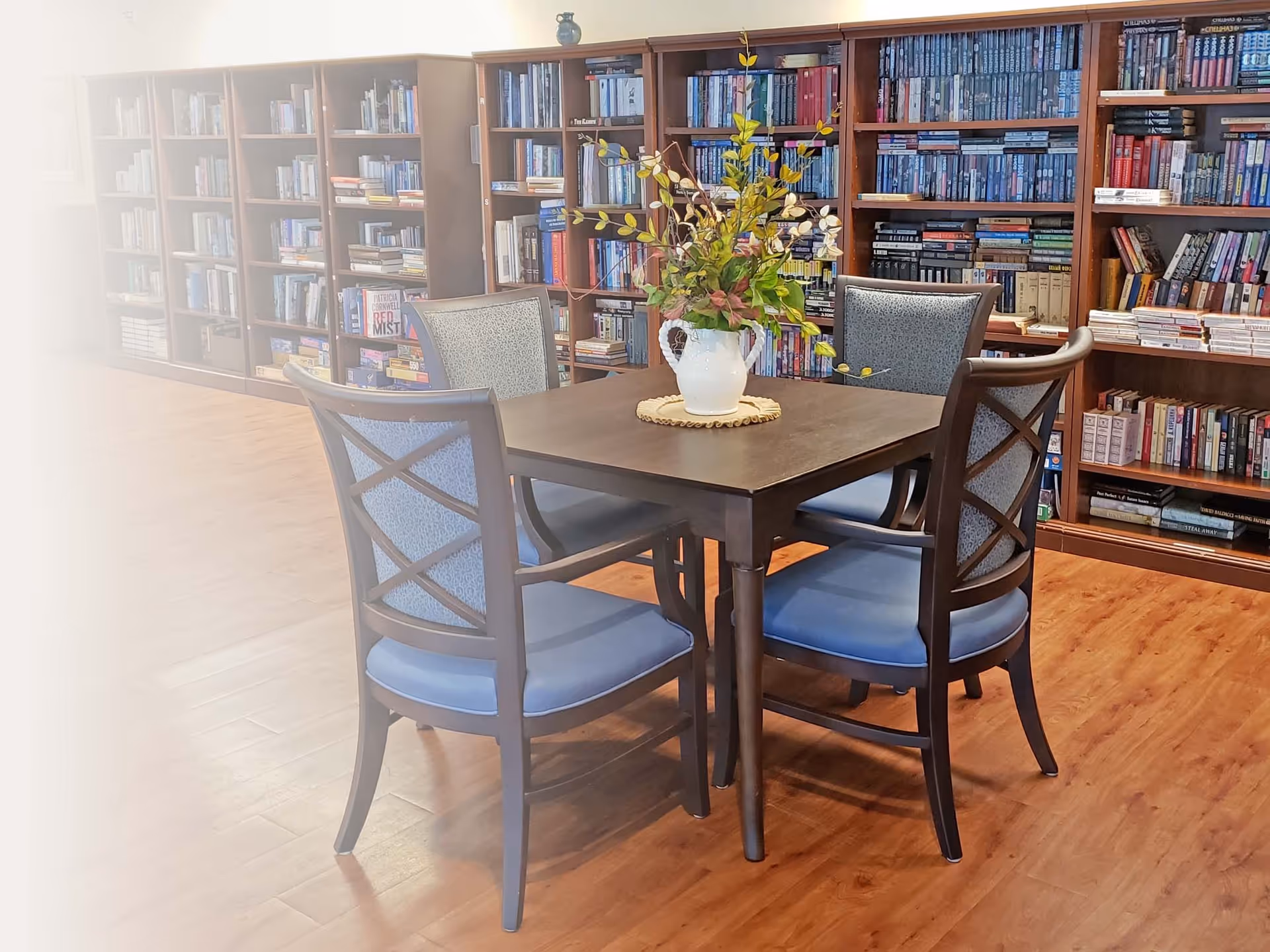 A library-style interior with a square wooden table surrounded by four blue-upholstered chairs and bookshelves lining the back wall.