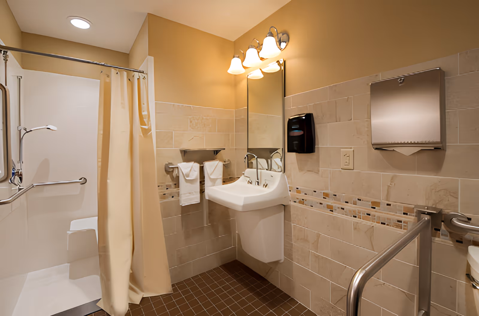 A clean and accessible bathroom with beige tiled walls and brown tiled floor. The bathroom features a white sink with a mirror above it, a wall-mounted soap dispenser, a paper towel dispenser, and two towel racks with white towels. There is a shower area with a beige curtain, grab bars, and a built-in seat. The lighting consists of three wall-mounted lamps above the mirror.