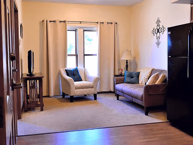 A cozy living room with an upholstered armchair and sofa, a side table with lamp, a TV, and a curtained window.