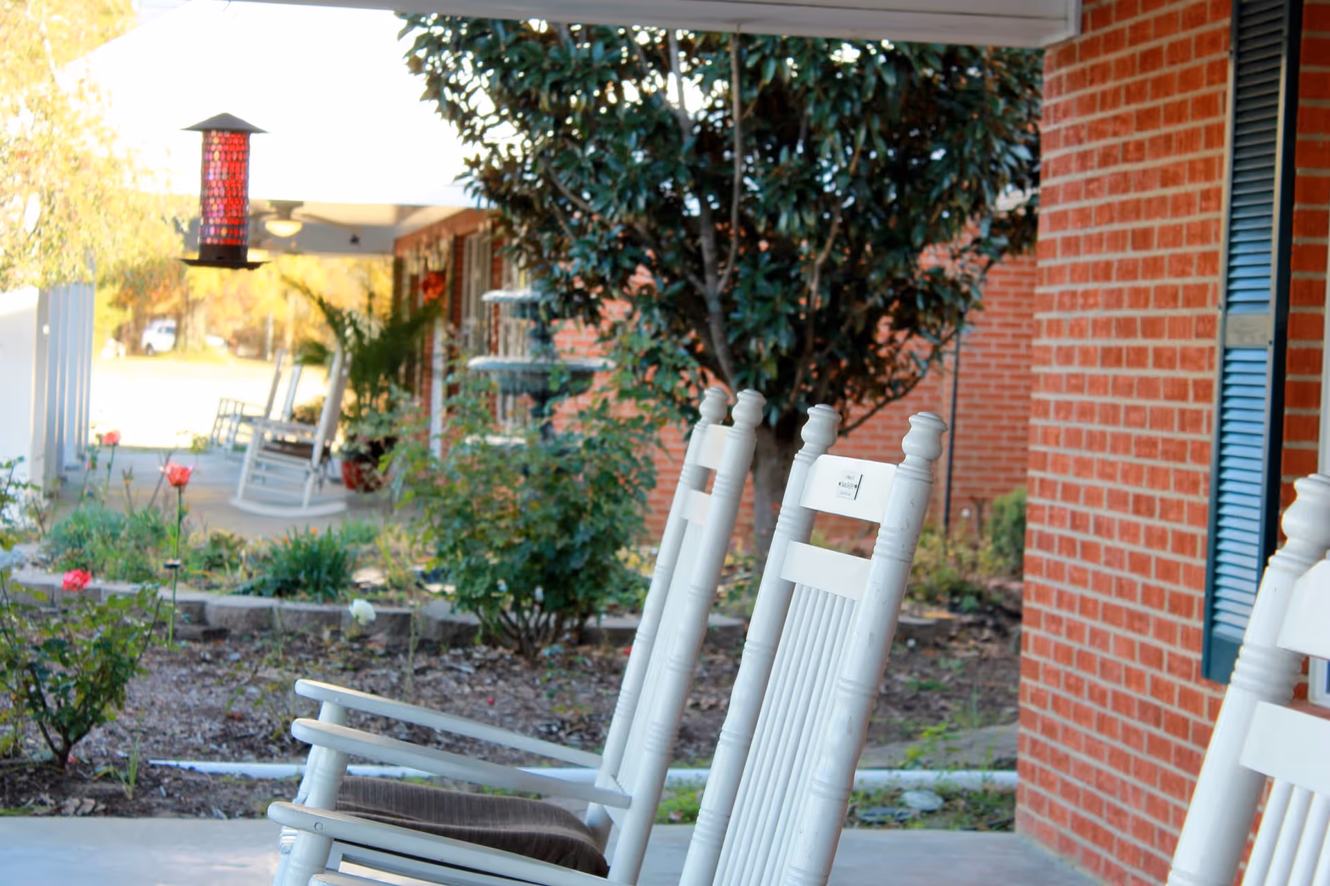 A view of white rocking chairs on a porch, surrounded by a garden with flowers and greenery, and a brick building in the background.