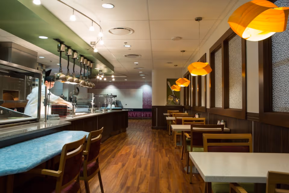 Interior dining room/cafeteria with a service counter and chef on the left and rows of tables under orange pendant lights on the right.