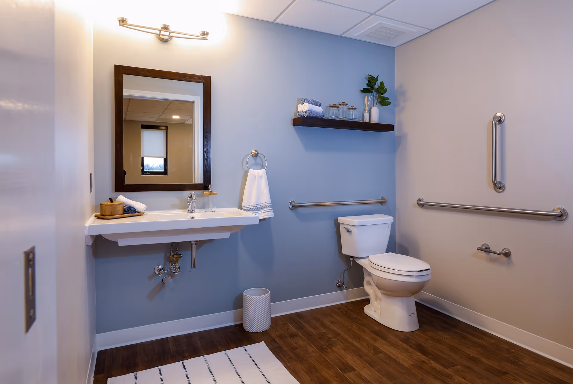 Accessible bathroom with sink, mirror, toilet, and grab bars on blue walls.