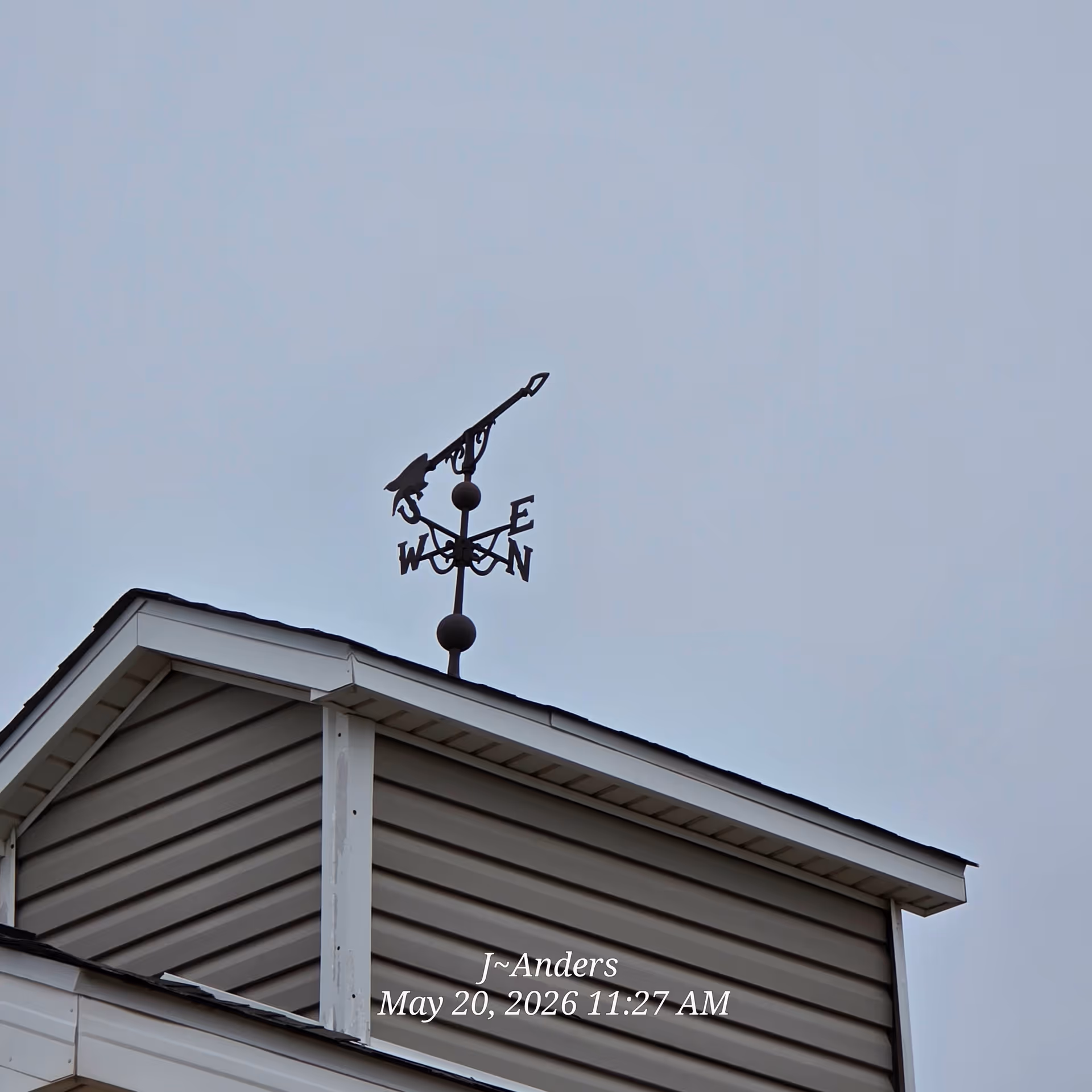 Weather vane atop a beige-siding roof against a pale blue sky.