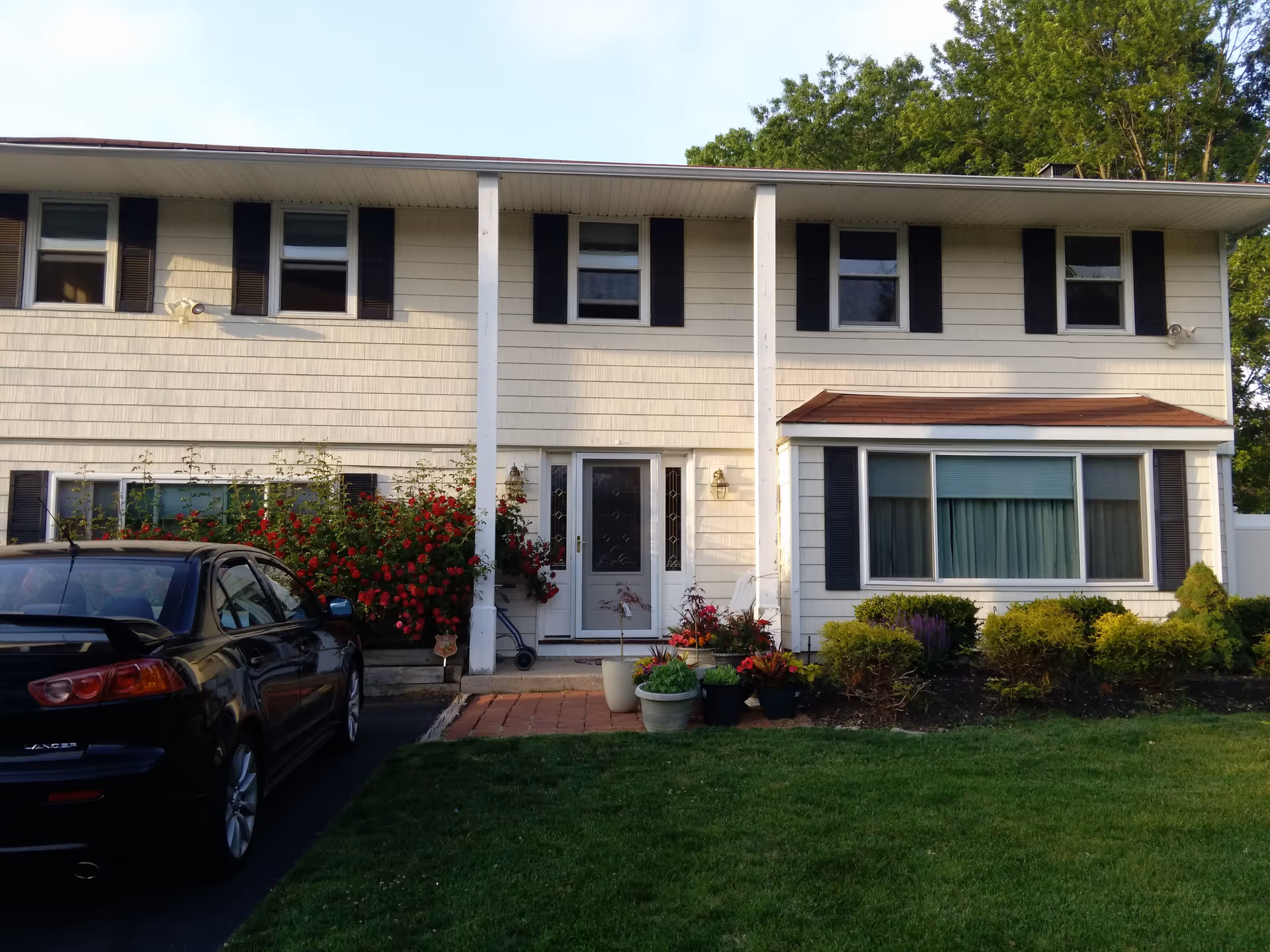 Front exterior of a two-story suburban house with a lawn, shrubs, potted plants, and a black car parked in the driveway.