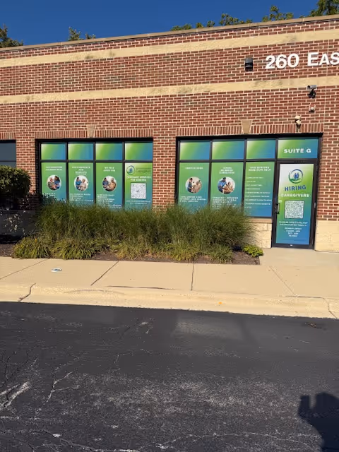 Brick storefront with green posters on windows, a door labeled with hiring signs, and tall bushes.