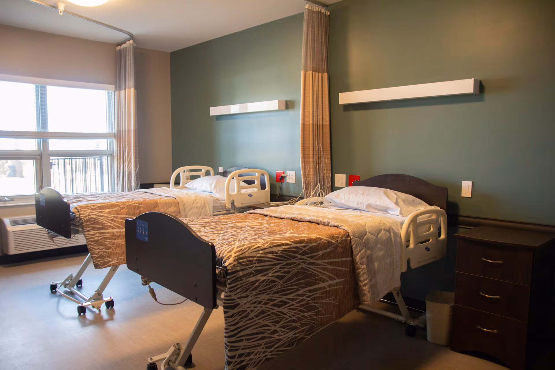 Two adjustable hospital-style beds with patterned covers in a shared senior living bedroom with a green accent wall and a window.