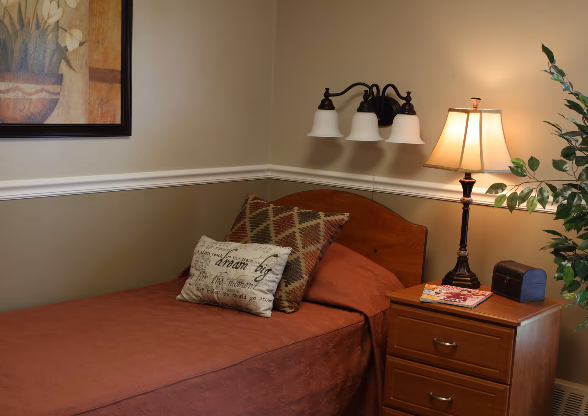 A cozy bedroom corner featuring a single bed with a rust-colored bedspread and two decorative pillows, one with a geometric pattern and the other with inspirational text. Above the bed is a three-light wall sconce, and next to the bed is a wooden nightstand with a table lamp, a small decorative chest, and some magazines. A framed floral painting hangs on the wall, and a green leafy plant is partially visible on the right side.