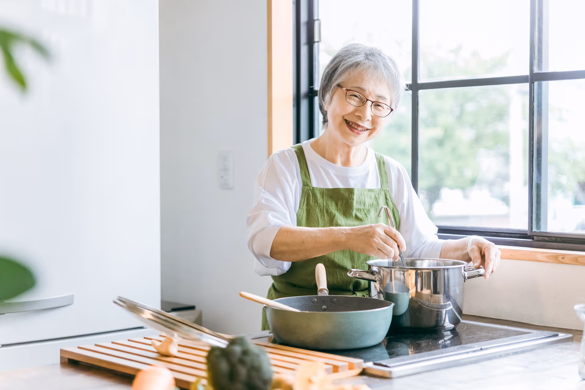 Smiling elderly woman in a green apron cooks at a stove in a bright kitchen.