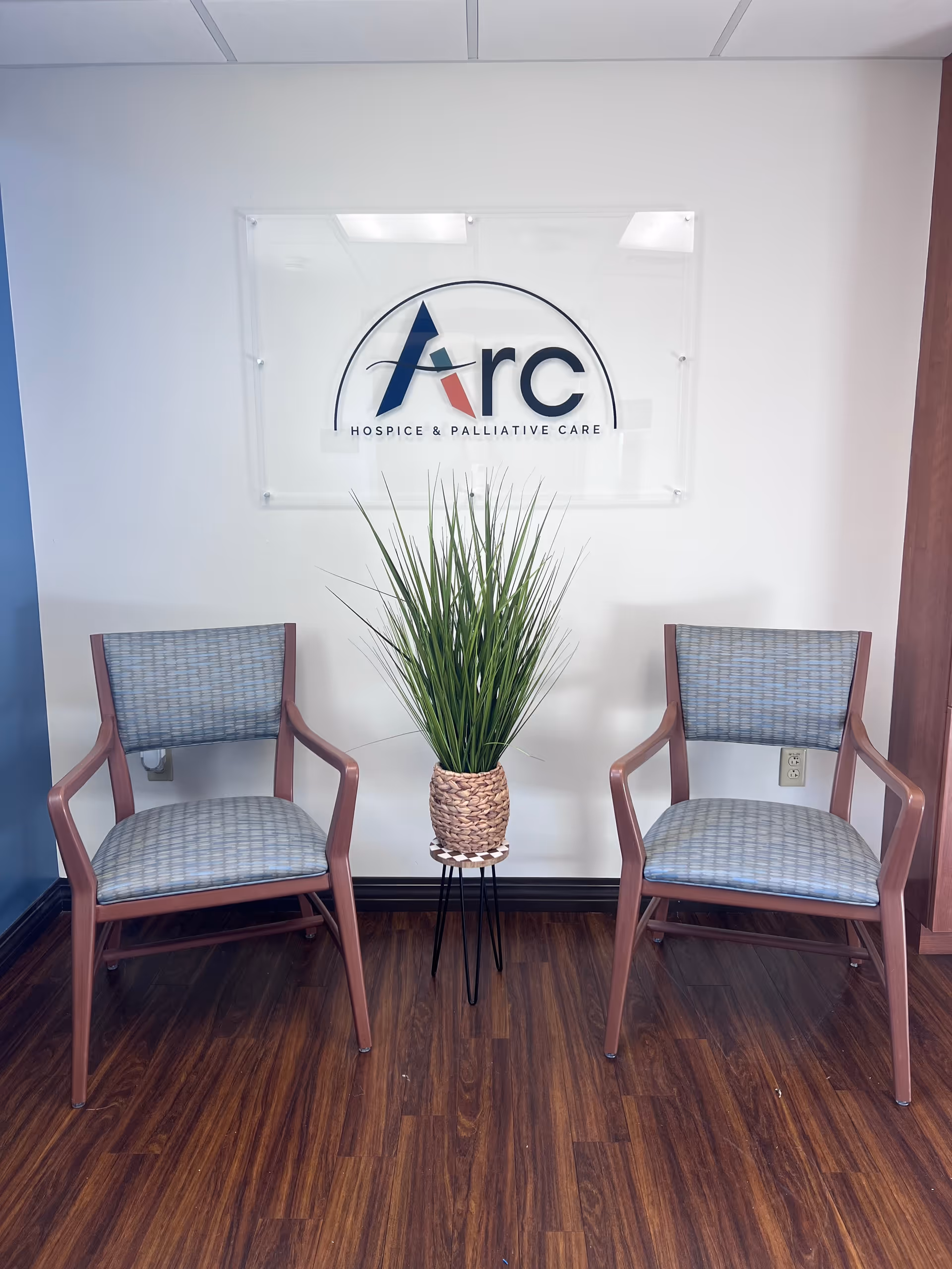 Two upholstered chairs flank a potted plant beneath Arc Hospice & Palliative Care sign.