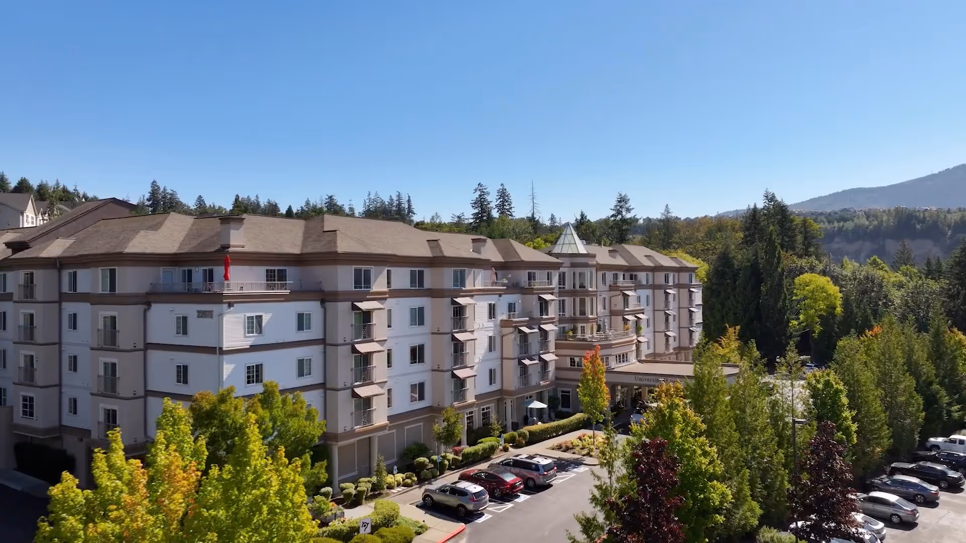 Exterior view of a multi-story senior living facility building surrounded by trees and a parking lot with several cars parked. The building has balconies and a peaked roof section at the entrance. The sky is clear and blue.