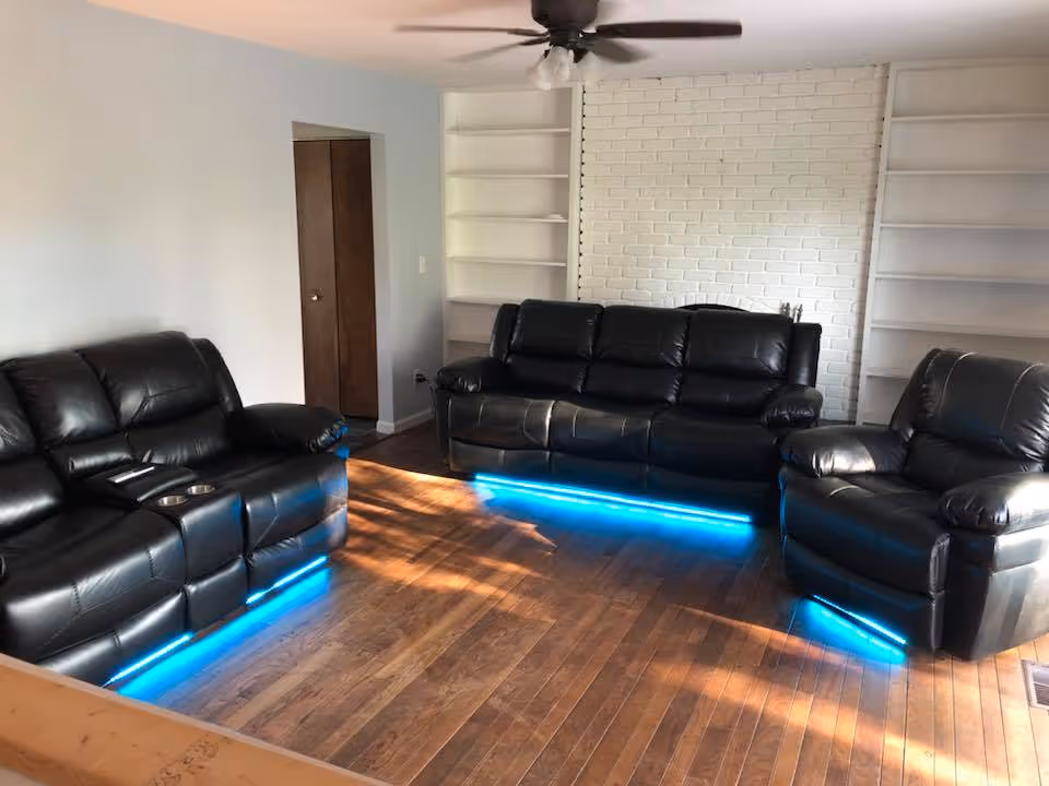 A living room with three black leather reclining sofas arranged around a wooden floor. The sofas have blue LED lights underneath them. There is a white brick wall with built-in shelves on either side and a ceiling fan above.