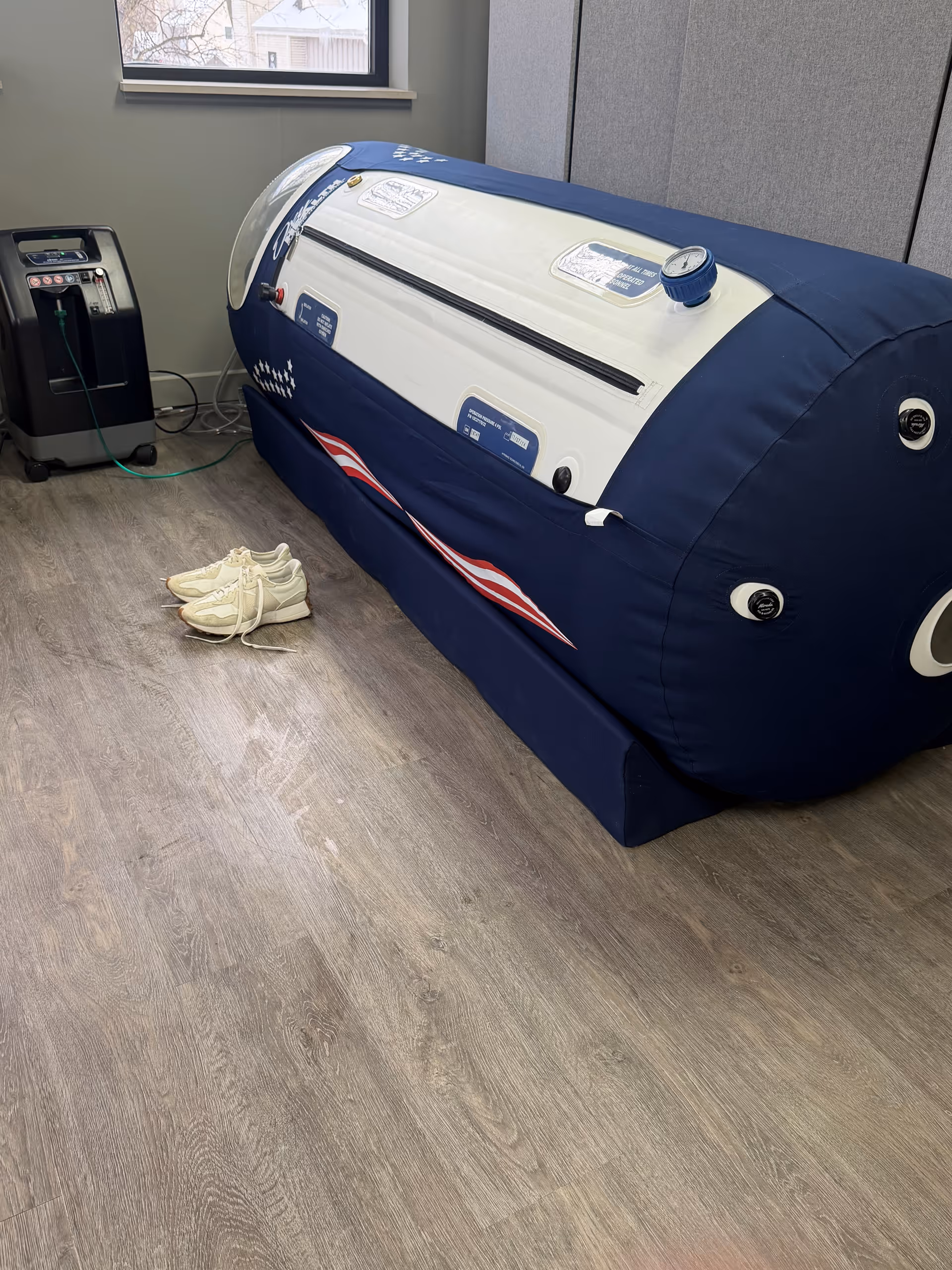 Blue cylindrical medical chamber in a clinical room, with shoes on wood-like floor beside a machine.