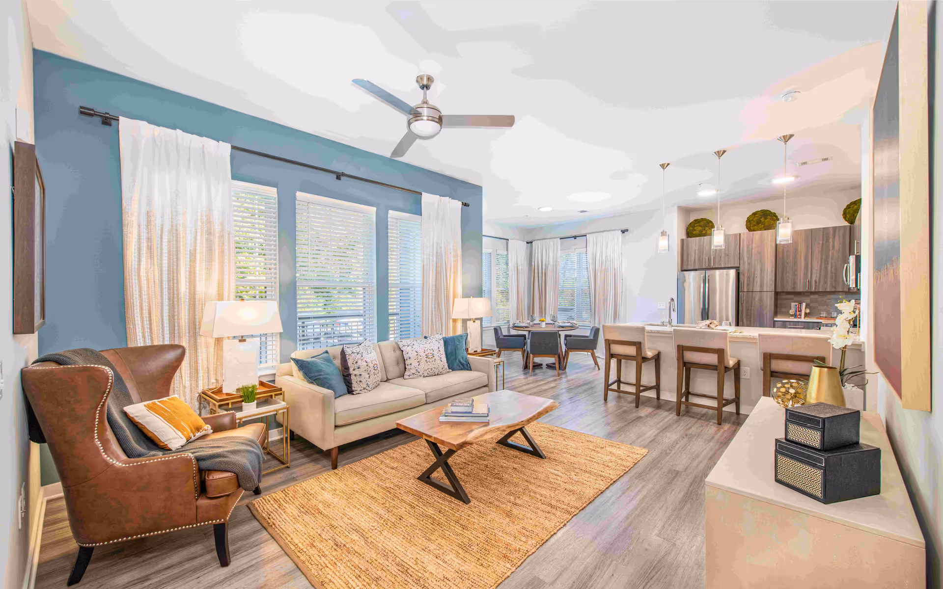 Bright open-plan living room and kitchen with a sofa, armchair, coffee table, dining area, and bar stools.
