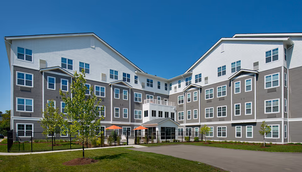 Exterior view of a modern four-story senior living facility building with gray and white siding, multiple windows, a fenced patio area with orange umbrellas, small trees, and a clear blue sky.