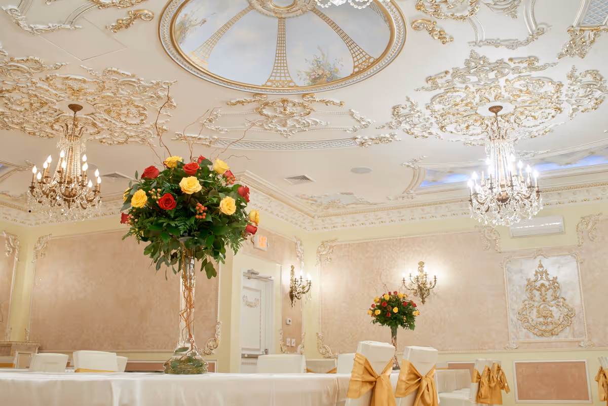 Ornate banquet hall with chandeliers, floral centerpieces, and gold-sashed chairs.