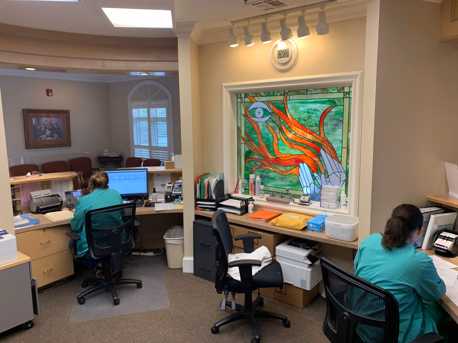 Two healthcare workers sit at desks with computers in a clinic reception area.