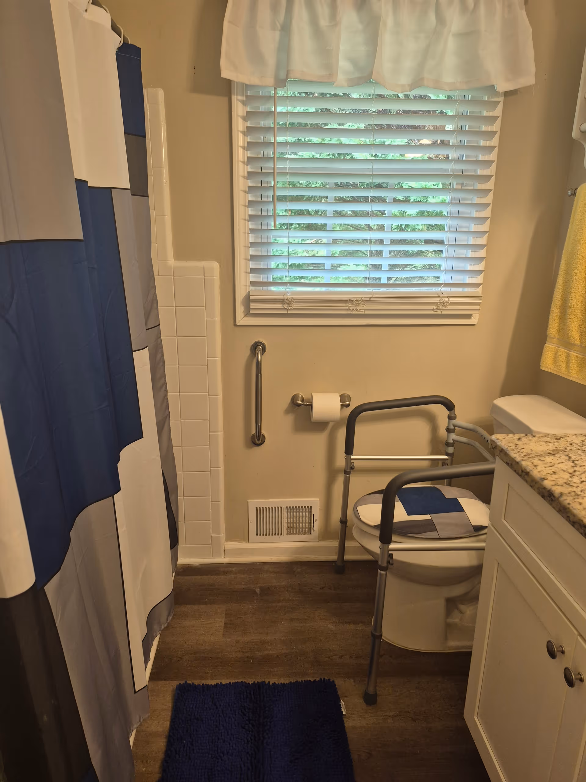 Small bathroom with shower curtain, window blinds, walker, toilet, and blue rug.