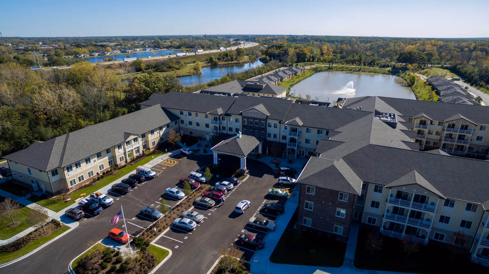 Aerial view of a multi-building senior living complex with a parking lot, entrance canopy and a pond with a fountain.