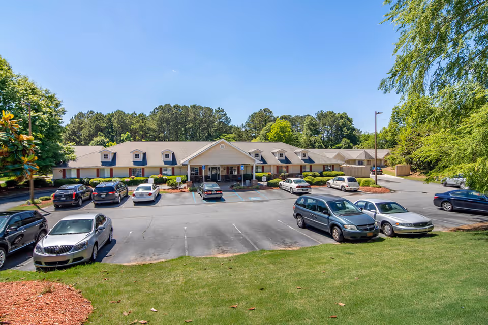 Exterior view of Bellevue Village at Neese Road senior living facility in Woodstock, showing a single-story building with a covered entrance, surrounded by trees and a parking lot with several parked cars under a clear blue sky.