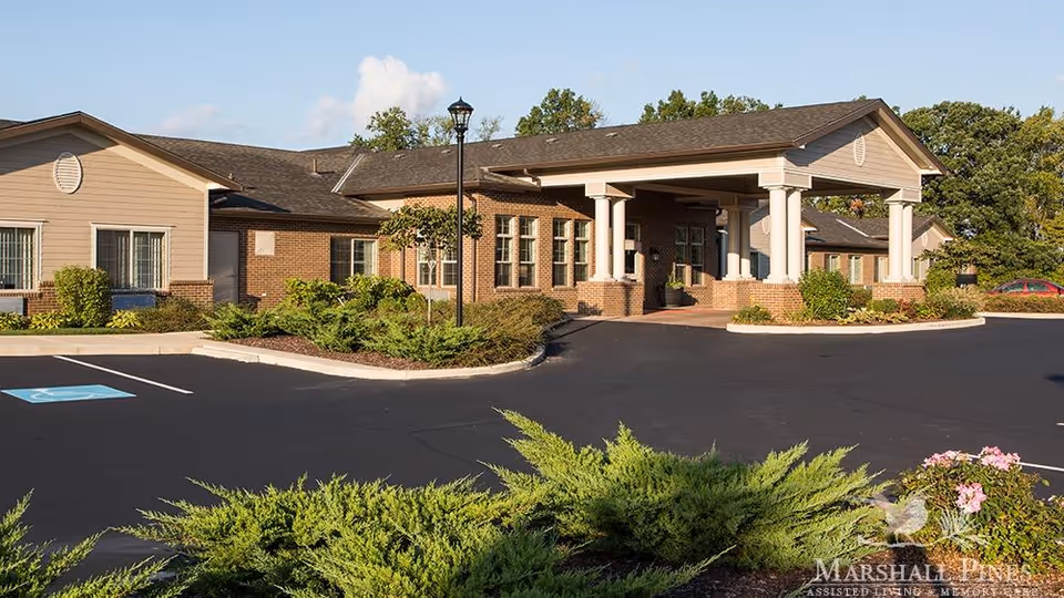 Exterior view of Marshall Pines Assisted Living & Memory Care building with a covered entrance supported by white columns, surrounded by landscaped bushes and trees, and a parking lot with a handicapped parking space.