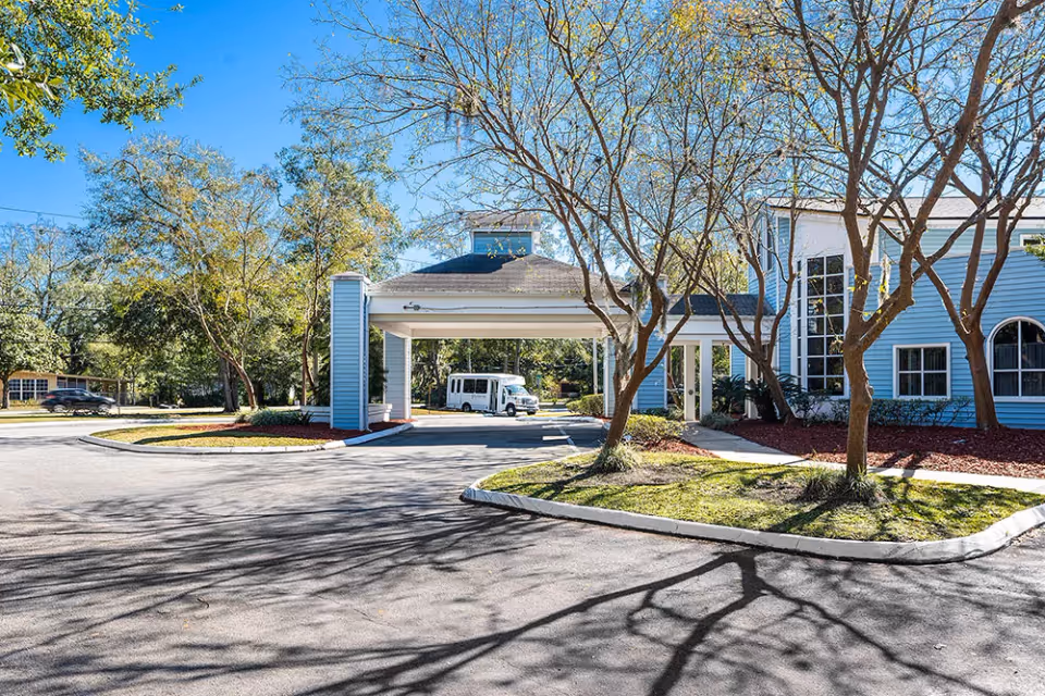 Exterior view of Fouraker Hills Rehab & Nursing Center showing a driveway with a covered entrance, a white shuttle bus parked under the entrance, and a blue building with large windows surrounded by trees and landscaping under a clear blue sky.