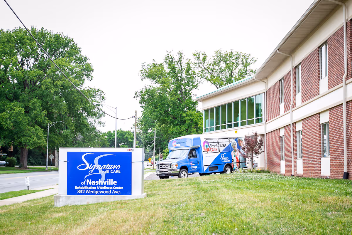 Exterior view of a brick senior living facility, sign, and a blue van parked outside.