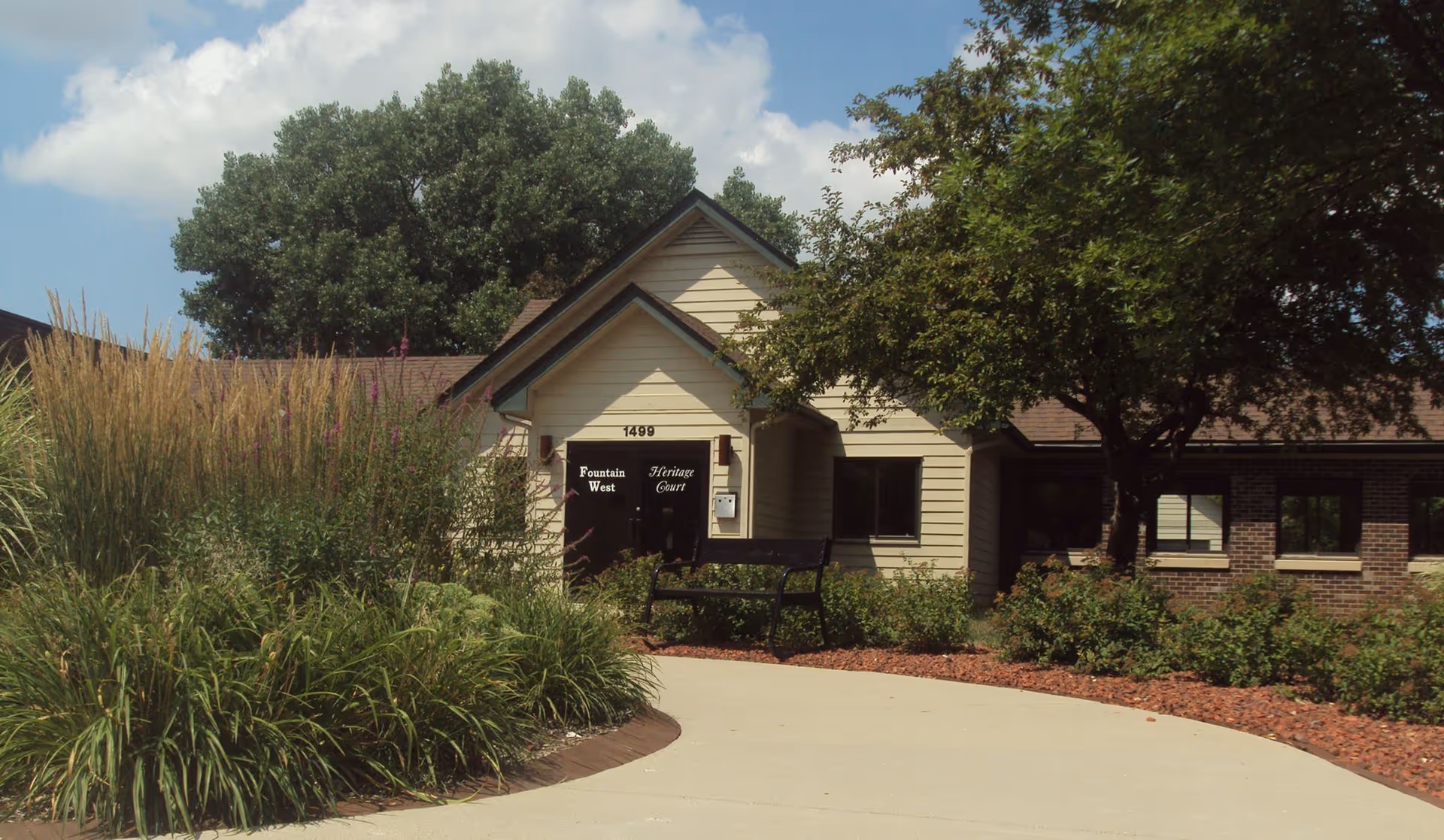 Front entrance of a single-story care building with a curved walkway, landscaping, bench, and signage by the door.