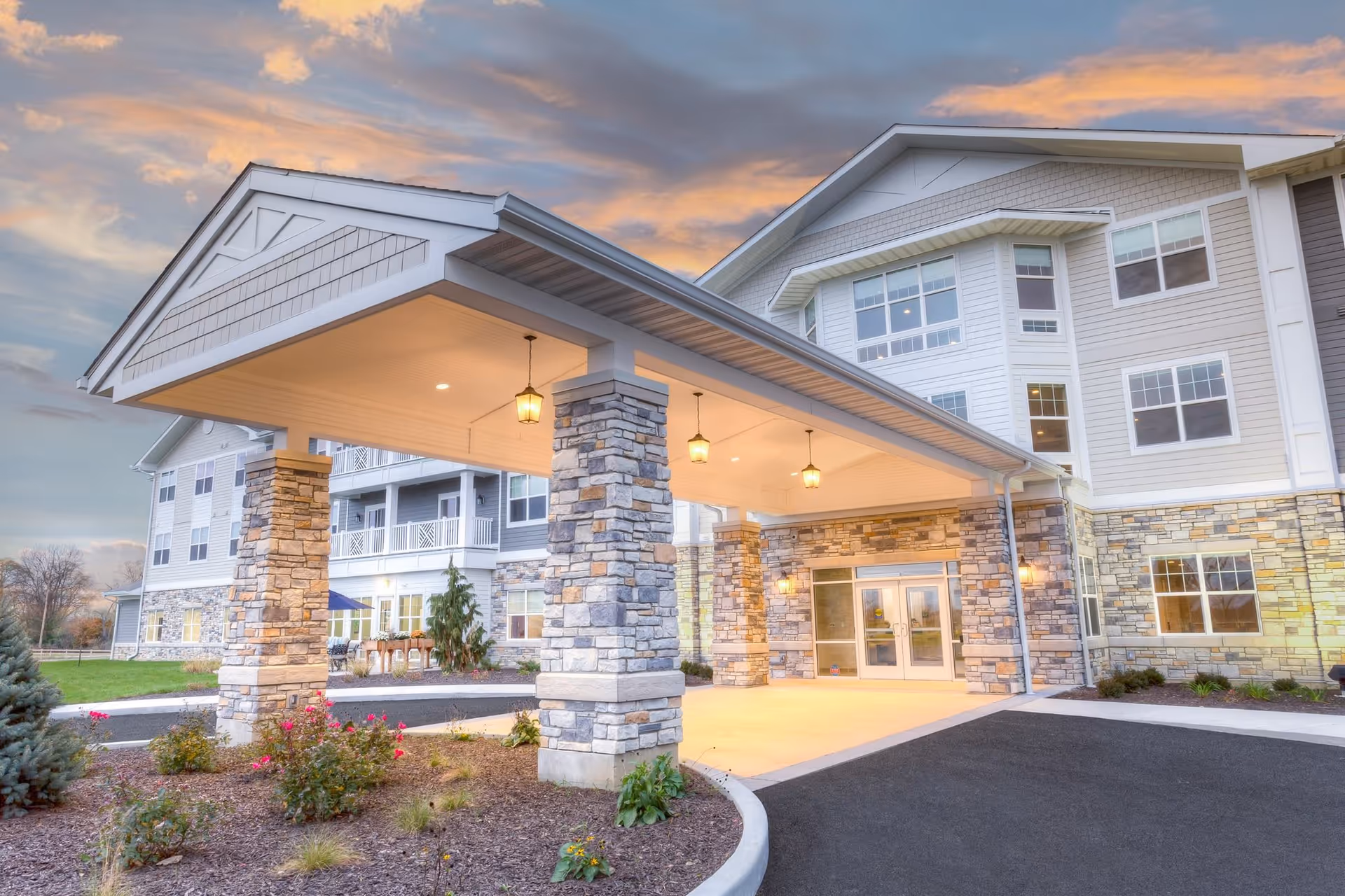 Entrance of a senior living facility named Residences at Coffee Creek during sunset, featuring a covered drop-off area supported by stone pillars, well-maintained landscaping with flowers and shrubs, and a multi-story building with numerous windows in the background.