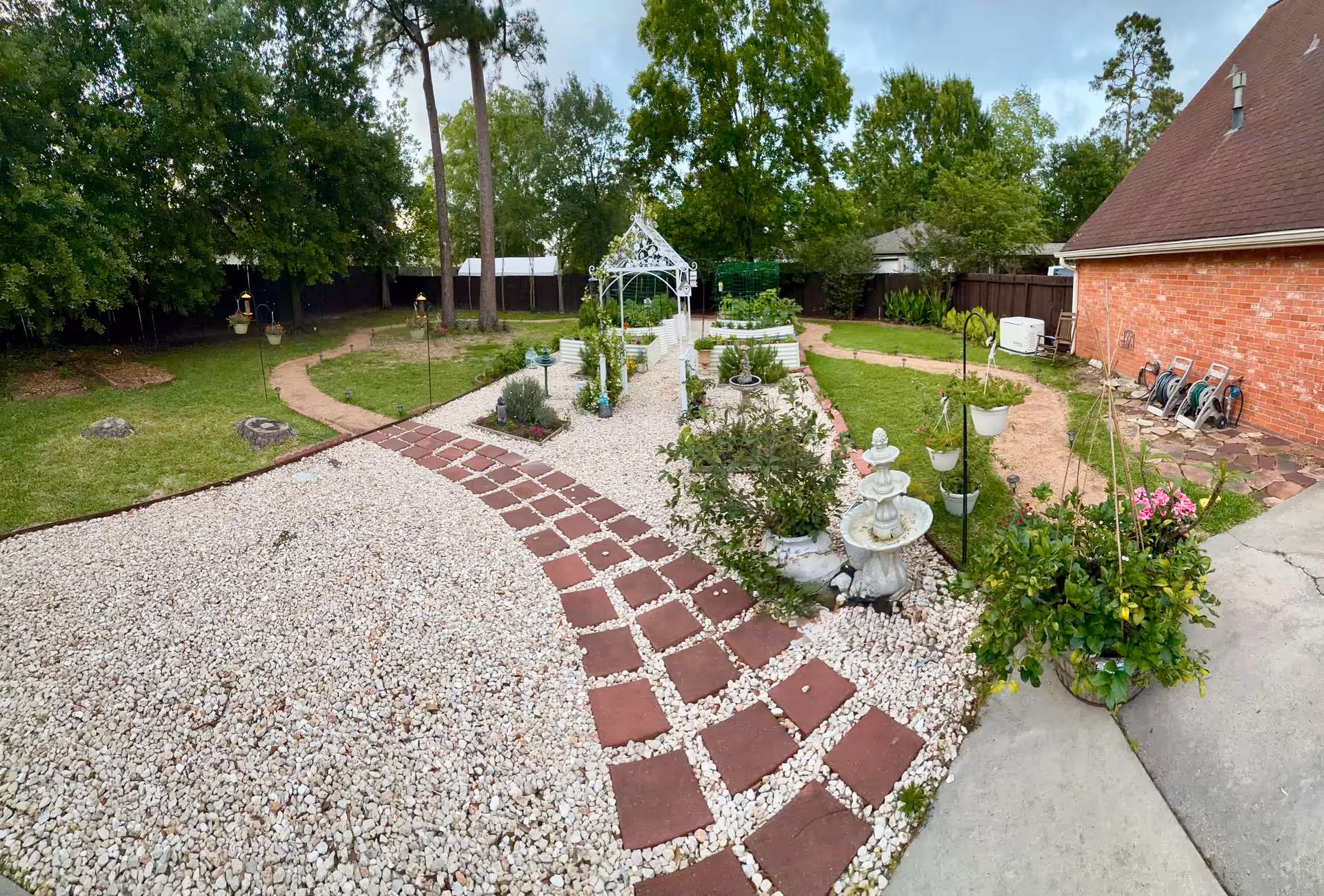 Extensive garden courtyard with gravel paths, a gazebo, fountain, and brick walkway beside a brick building.