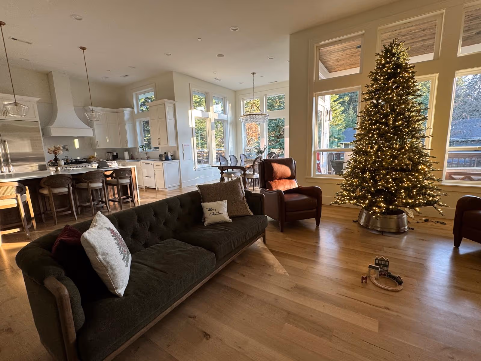Open living area with Christmas tree, sofa, chairs, and bright kitchen in background