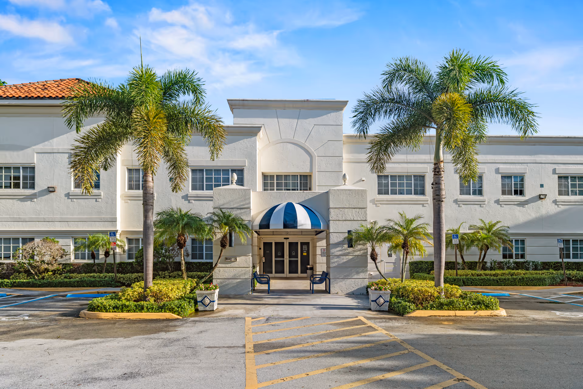 Exterior view of a white senior living facility entrance with palm trees and blue awning.
