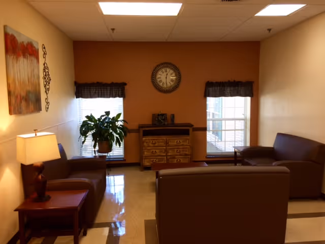 A cozy living room area with brown leather sofas arranged around a small wooden chest of drawers. Two windows with dark valances flank the chest, and a round clock hangs on the wall above it. A potted plant is placed near one window, and a table lamp sits on a wooden side table next to one of the sofas. The walls are painted in warm tones, and a piece of artwork is hung on the left wall.
