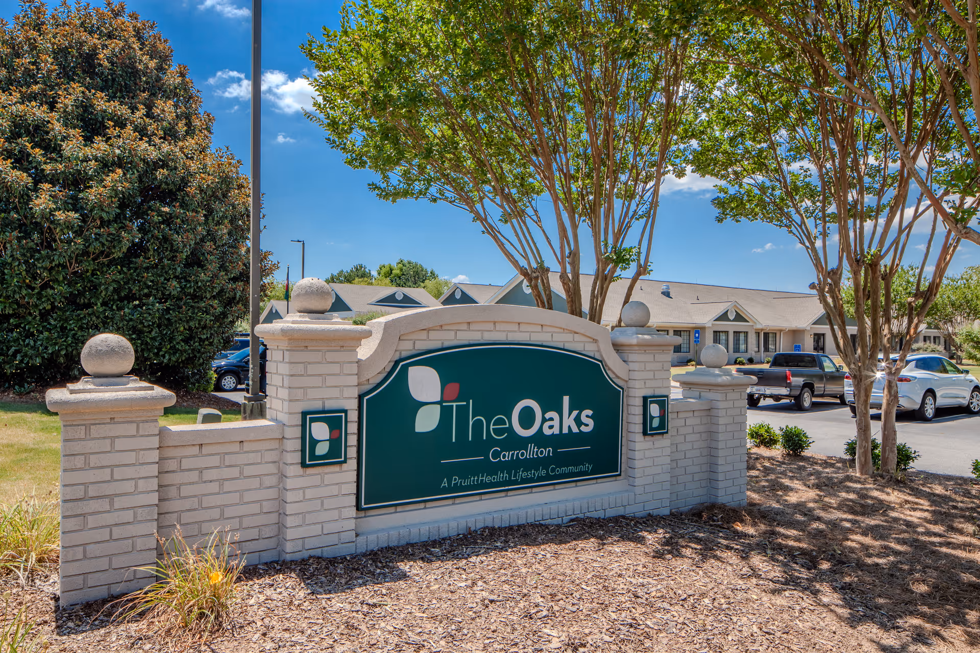 Exterior sign for The Oaks Carrollton with trees, mulch, and parked cars.