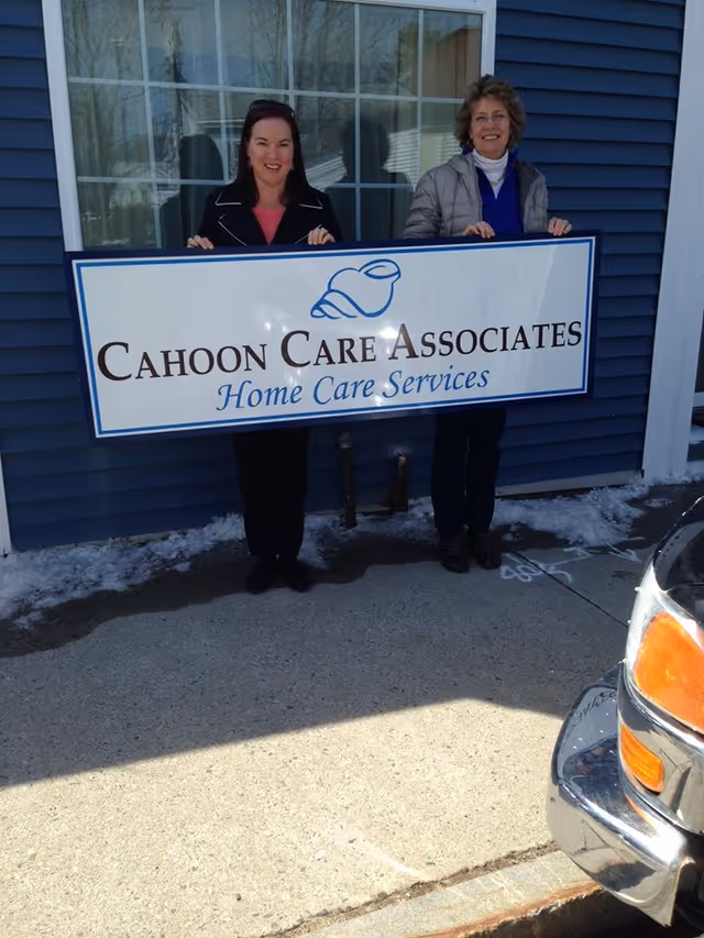 Two women hold a Cahoon Care Associates sign outside a blue building.