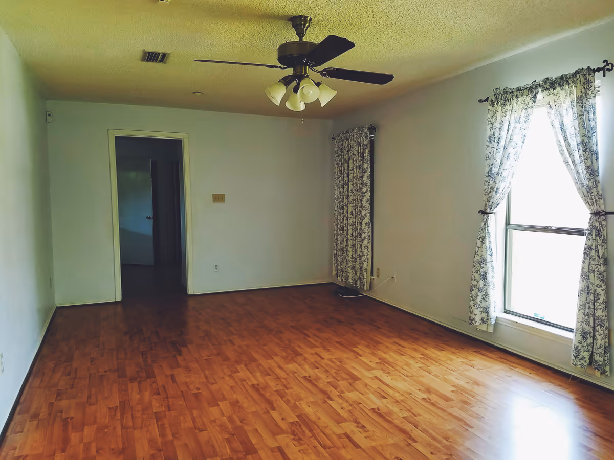 Empty room with wooden flooring, white walls, a ceiling fan with lights, and two windows with floral curtains letting in natural light. There is a doorway leading to another room.