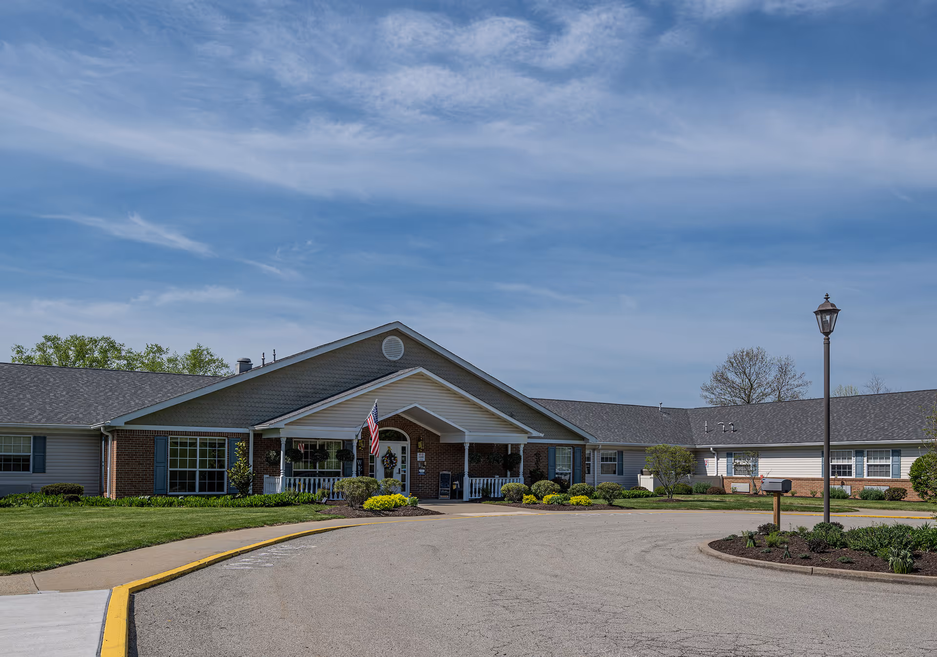Exterior view of a single-story senior living facility building with a covered entrance, an American flag, well-maintained landscaping, and a circular driveway under a partly cloudy blue sky.