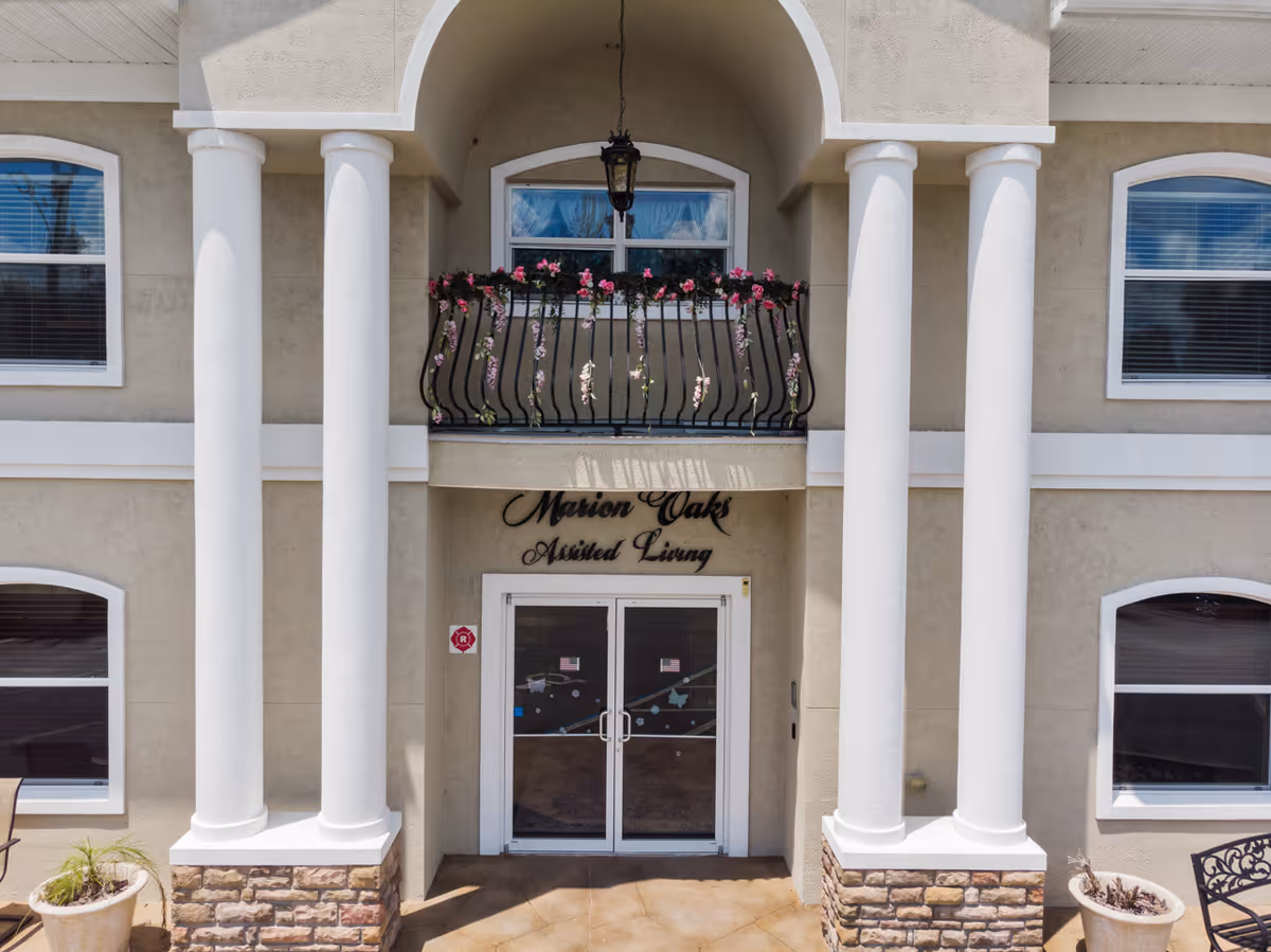Front entrance of Marion Oaks Assisted Living facility featuring a double glass door, four large white columns, a small balcony with a black wrought iron railing decorated with pink flowers, and beige exterior walls with white trim around windows.