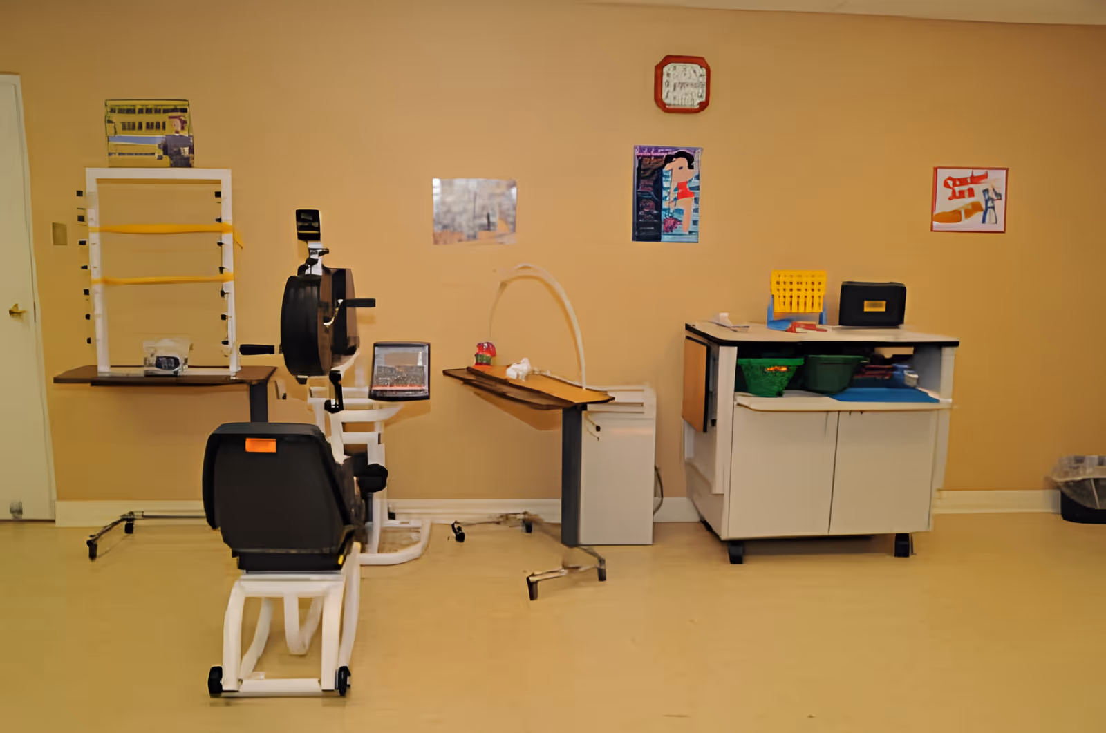 A therapy or rehabilitation room with exercise equipment including a stationary hand cycle machine, a table with therapy tools, and a cabinet with various supplies. The walls are beige with a few colorful posters and a clock hanging above.