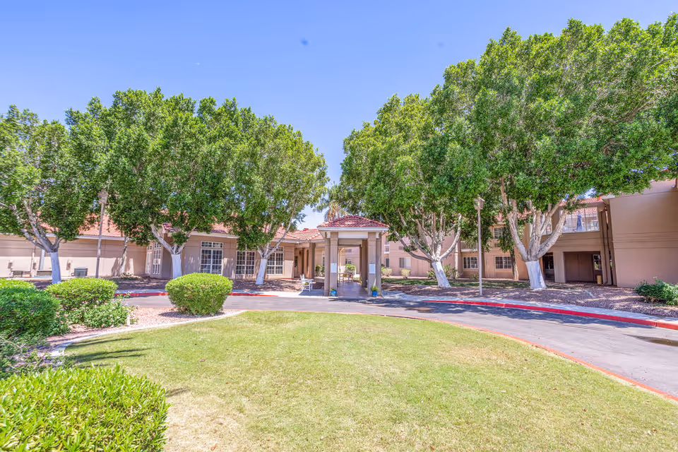 Front entrance of a single-story senior living building with a covered porte-cochère, surrounding trees, shrubs, and a circular driveway.