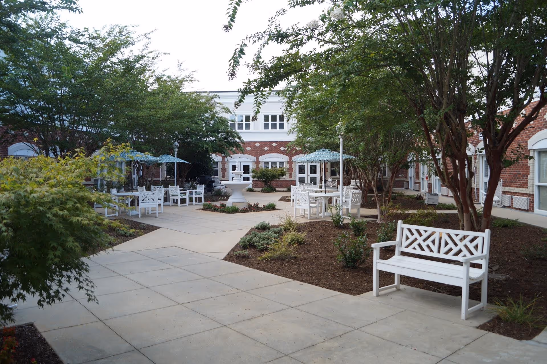 Outdoor courtyard area with white benches, tables with blue umbrellas, trees, and a white fountain in the center, surrounded by a red brick building with white trim windows.