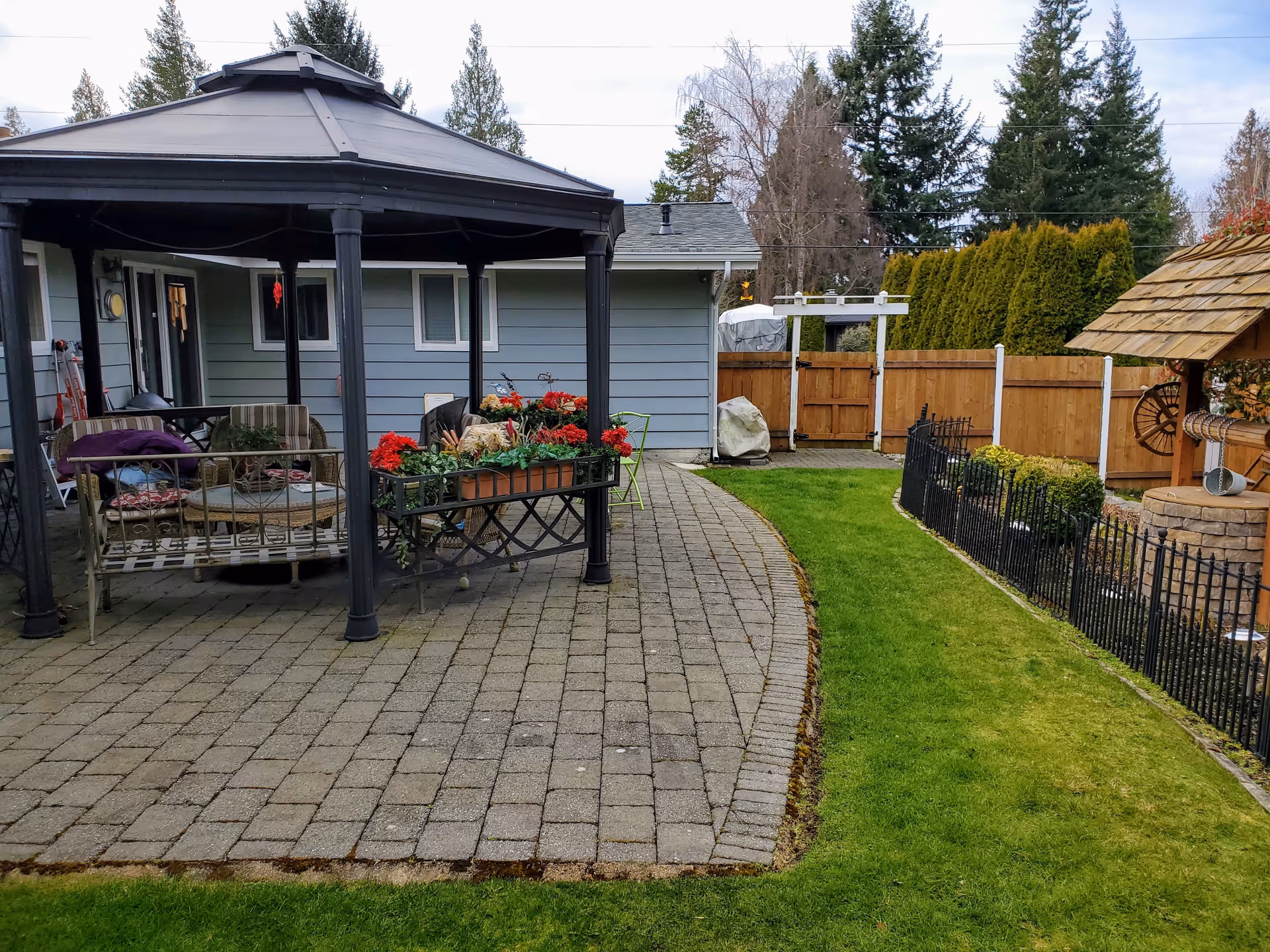 A backyard patio area with a black gazebo covering outdoor furniture including chairs and a table. There are flower boxes with red flowers on the patio. The patio is paved with stone tiles and is adjacent to a green lawn. A wooden fence and some trees are visible in the background, along with a small well-like structure with a shingled roof.